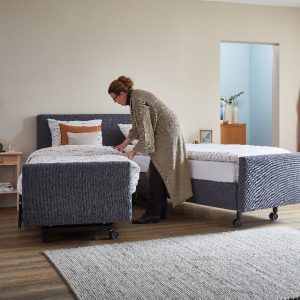 grijze boxspring met wielen welke uit elkaar is gereden, met een vrouw welke het bed opmaakt