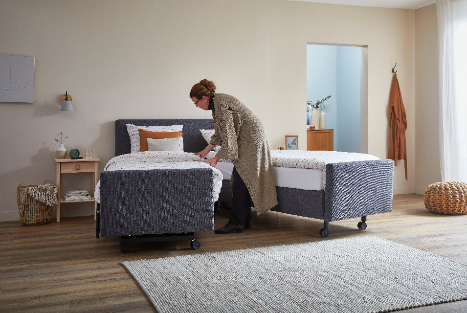 grijze boxspring met wielen welke uit elkaar is gereden, met een vrouw welke het bed opmaakt