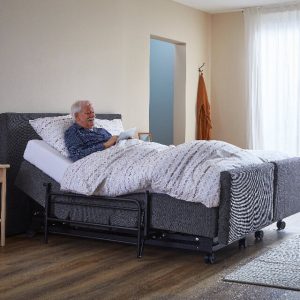 Grijs hoog laag senioren bed met uitval rek en een oudere man welke in bed ligt met tablet